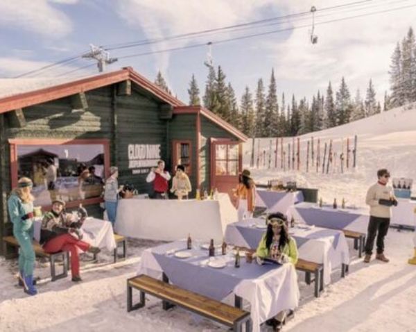 A group of diners enjoying meals at tables outside a ski lodge in a winter setting, surrounded by snow-covered mountains.