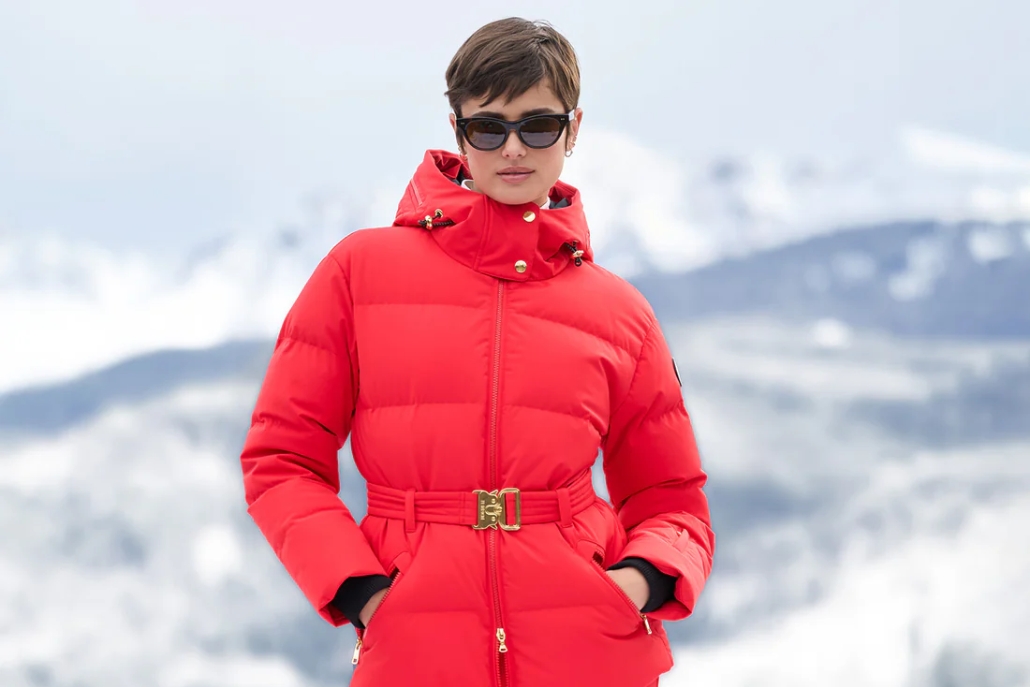 A woman in a stylish red belted coat and sunglasses stands on a snowy mountain, representing Aspen's winter fashion trends.
