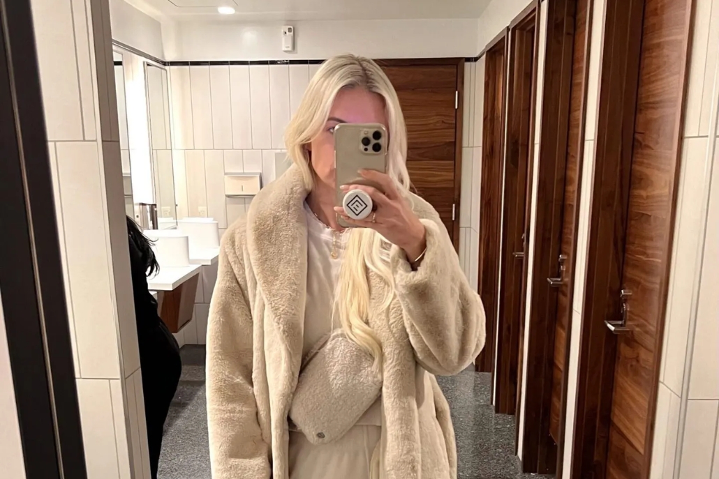 A woman in a stylish white coat poses for a selfie, reflecting her unique fashion.