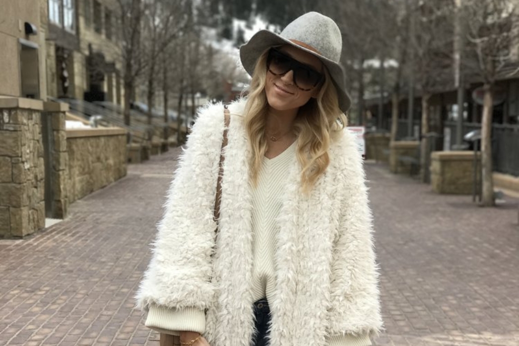 Blonde woman wearing a white fuzzy coat and hat, embodying Aspen Style in a snowy environment.