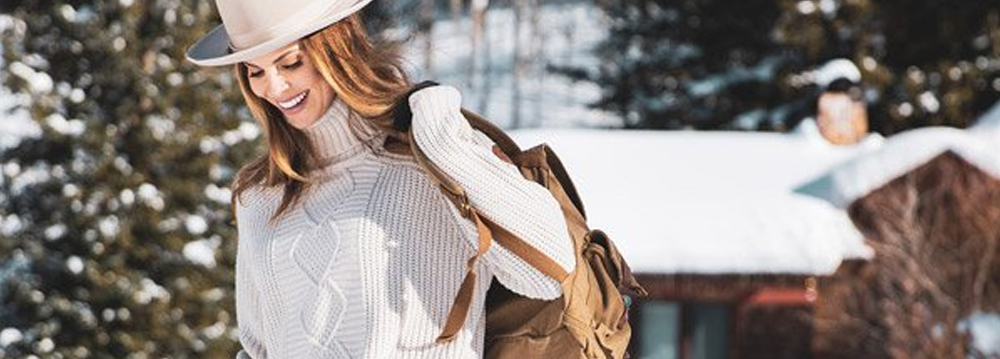 A woman wearing a hat and sweater walks in the snow, showcasing the charm of Aspen Style.