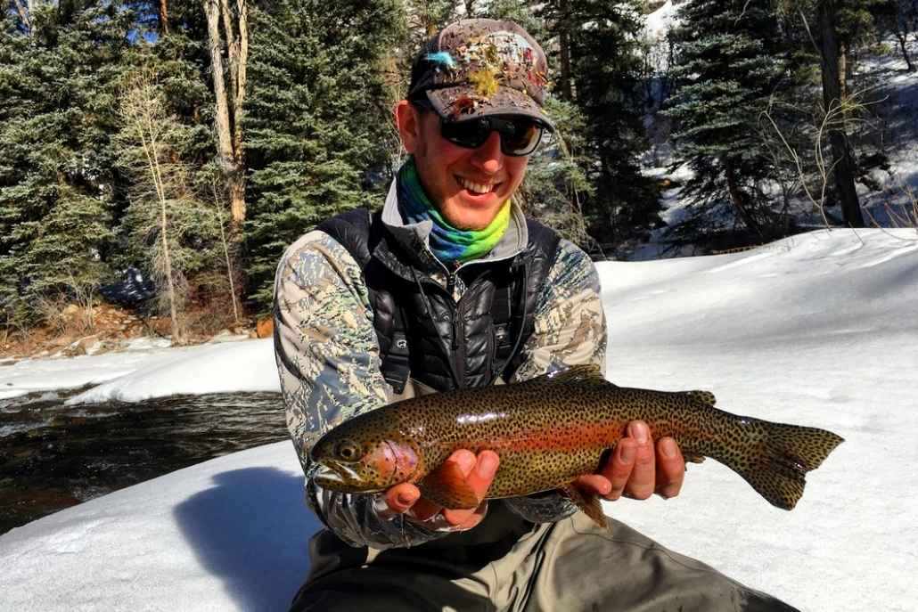 A man proudly displays a rainbow trout in the snow, showcasing his fishing success