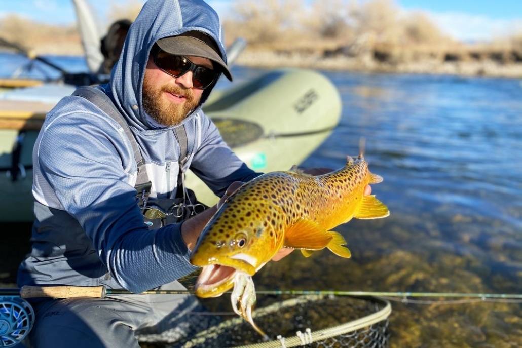 A man in a hoodie proudly holds a brown trout, showcasing his successful catch