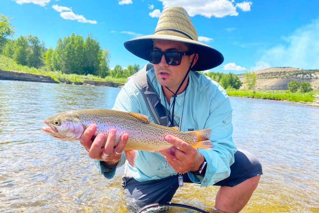 A man wearing a hat and sunglasses proudly holds a rainbow trout in Aspen River