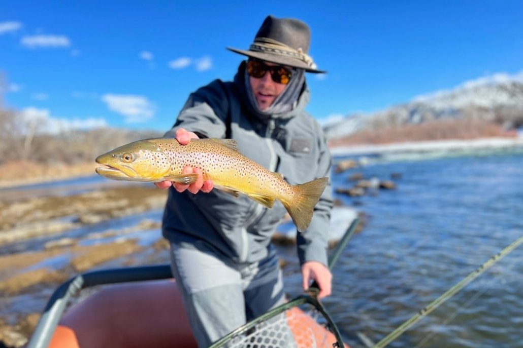 A man with sunglasses and a hat holds a fish, representing his experience in Aspen Fly Fishing.