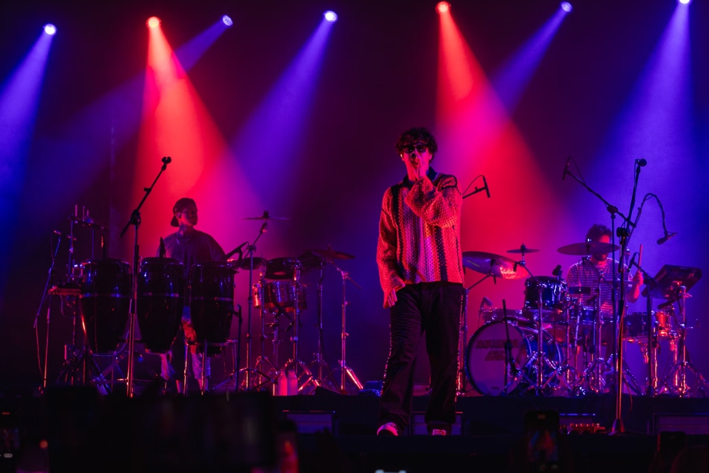 A musical group plays on stage, surrounded by bright red and blue lights, set against the backdrop of Aspen, Colorado.