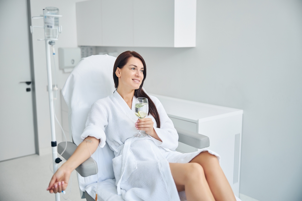 Woman in a white robe seated in a hospital chair with a glass of water, highlighting IV therapy services at The Fix, Aspen.