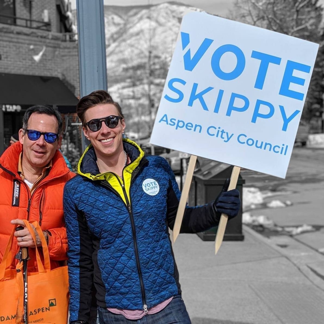 Two men stand together, holding a sign that encourages participation in the voting process.