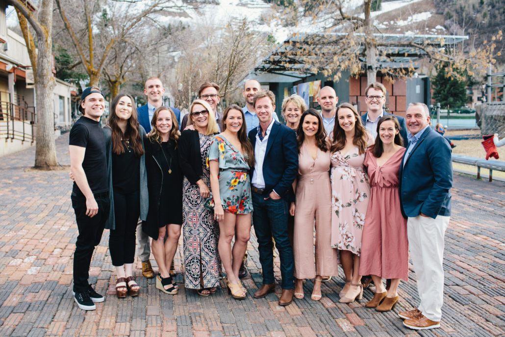 A group of family and friends of newlywed smiling and posing together in front of a large building during a sunny day.