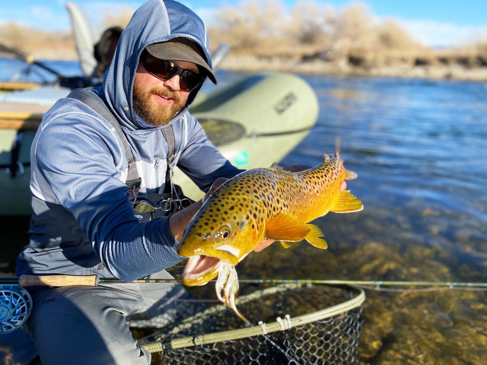 A man in a hoodie proudly holds a brown trout, showcasing his successful catch