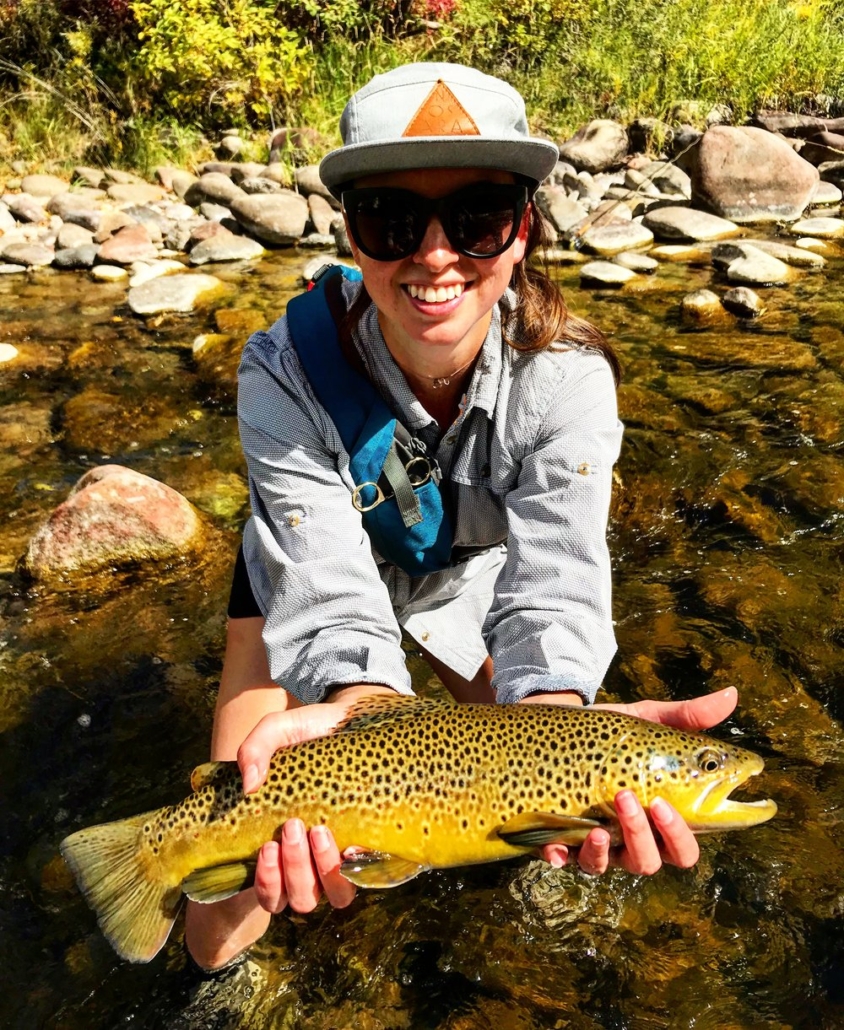 A woman stands in water, proudly holding a brown trout, showcasing her Aspen Fly Fishing success.