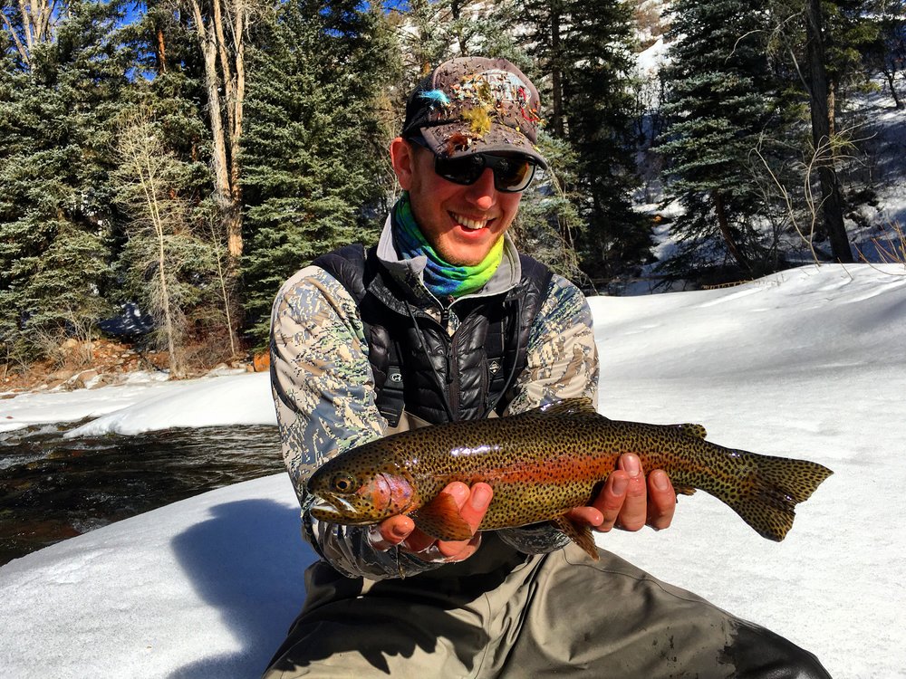 A man proudly displays a rainbow trout in the snow, showcasing his fishing success