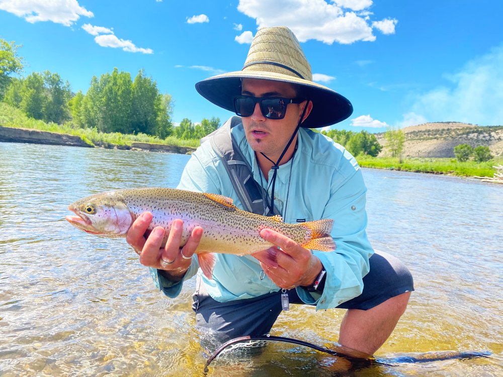 A man wearing a hat and sunglasses proudly holds a rainbow trout in Aspen River