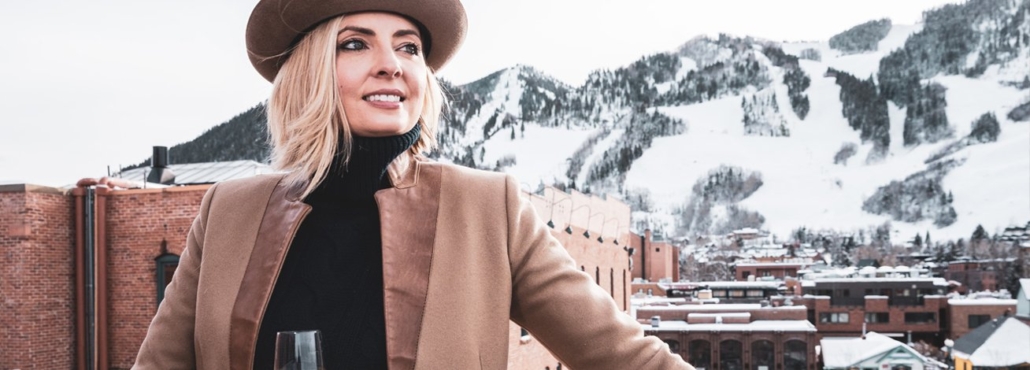 A woman in a hat and coat stands on a rooftop, overlooking mountains in the background.