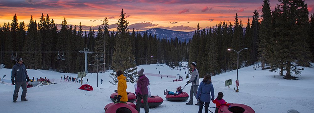 Groups of people enjoying snow tubing down a slope, surrounded by a picturesque sunset backdrop during Aspen Winter.