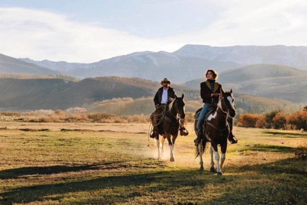 Two riders on horseback traverse a scenic mountain trail, surrounded by stunning peaks and greenery.