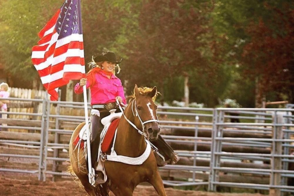 A woman on horseback holds an American flag, showcasing her spirit in a beautiful landscape.