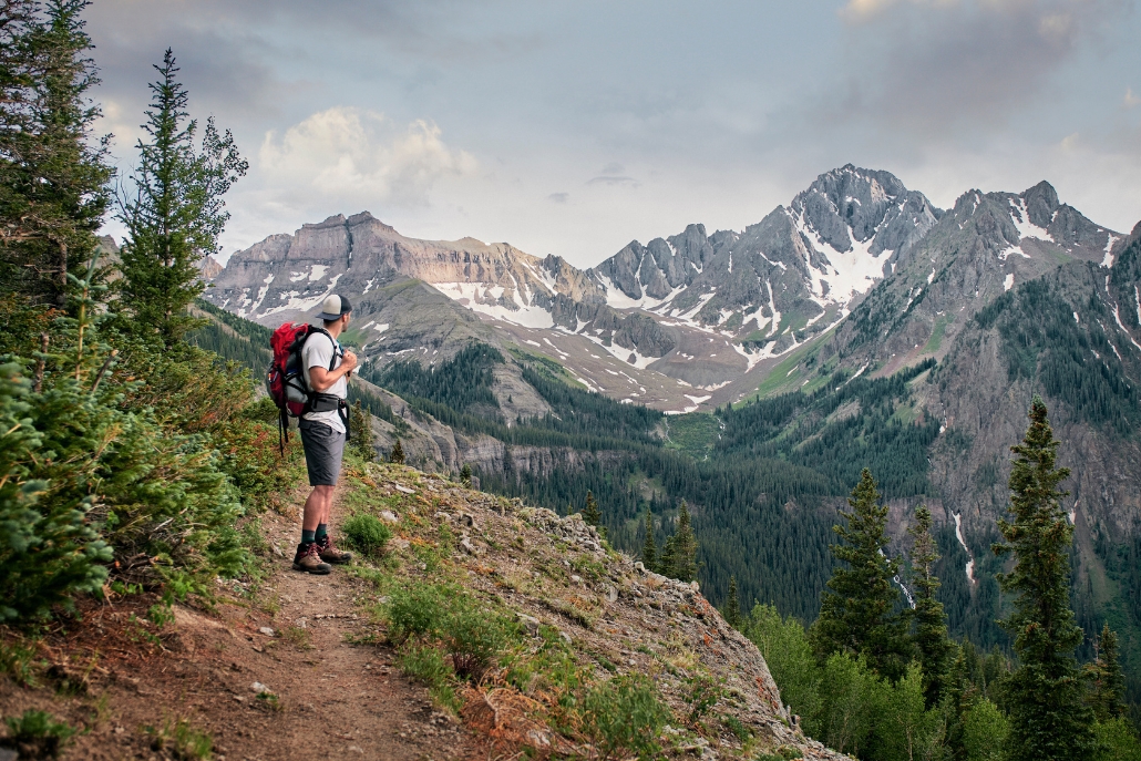 A man with a backpack stands on the Cathedral Lake Trail, surrounded by mountains and lush greenery.