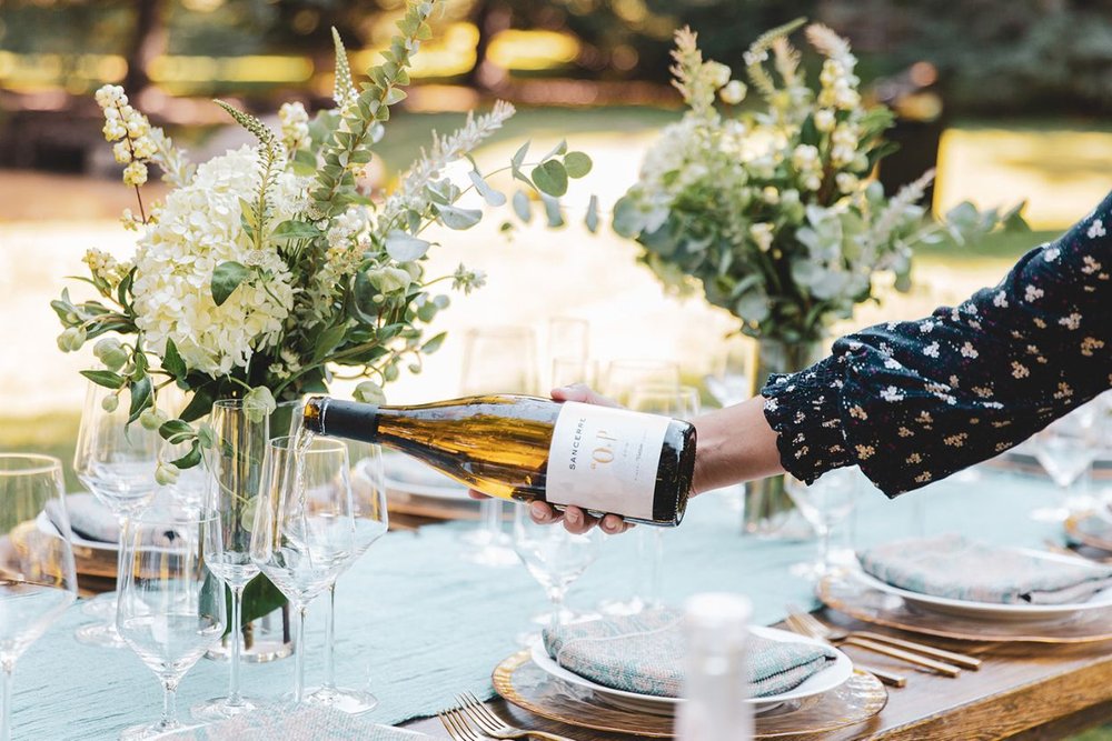 Elegant outdoor picnic setup in Aspen with wine pouring and lush floral decorations, set against a natural, green backdrop in July.