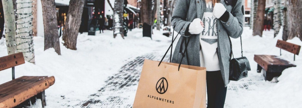 A woman joyfully holds a shopping bag while walking through the snow, embodying the spirit of Shopping in Aspen.