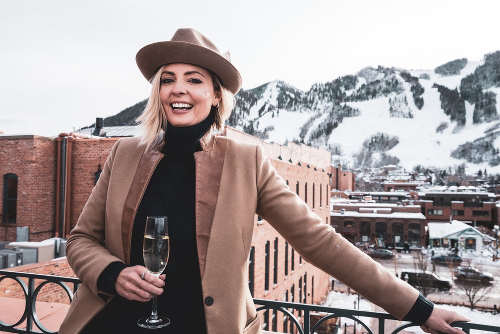 A woman wearing a fashionable hat and coat holds a glass of wine, exuding sophistication and relaxation.