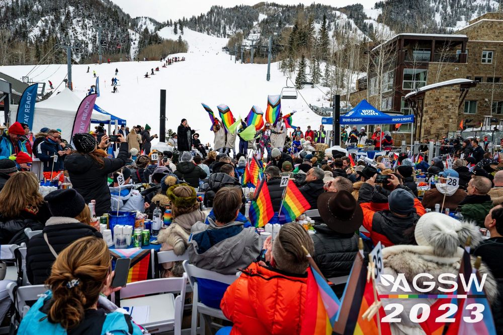 A lively crowd gathers at a ski resort, surrounded by pride flags fluttering in the winter breeze during Aspen Gay Ski Week