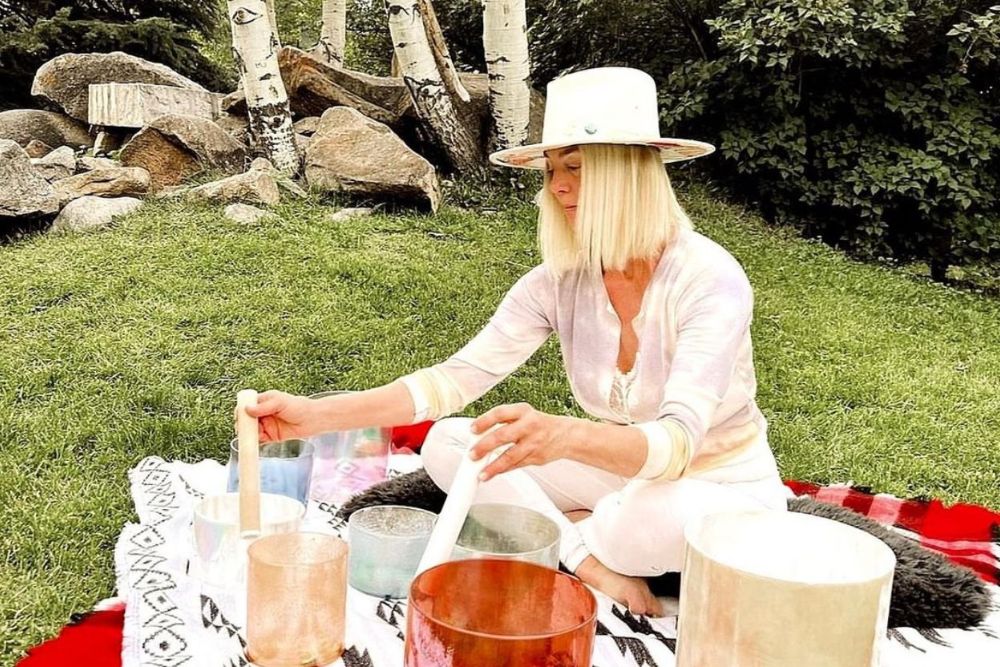 A woman in a hat relaxes on a blanket, performing Aspen Shakti, surrounded by a peaceful outdoor setting.