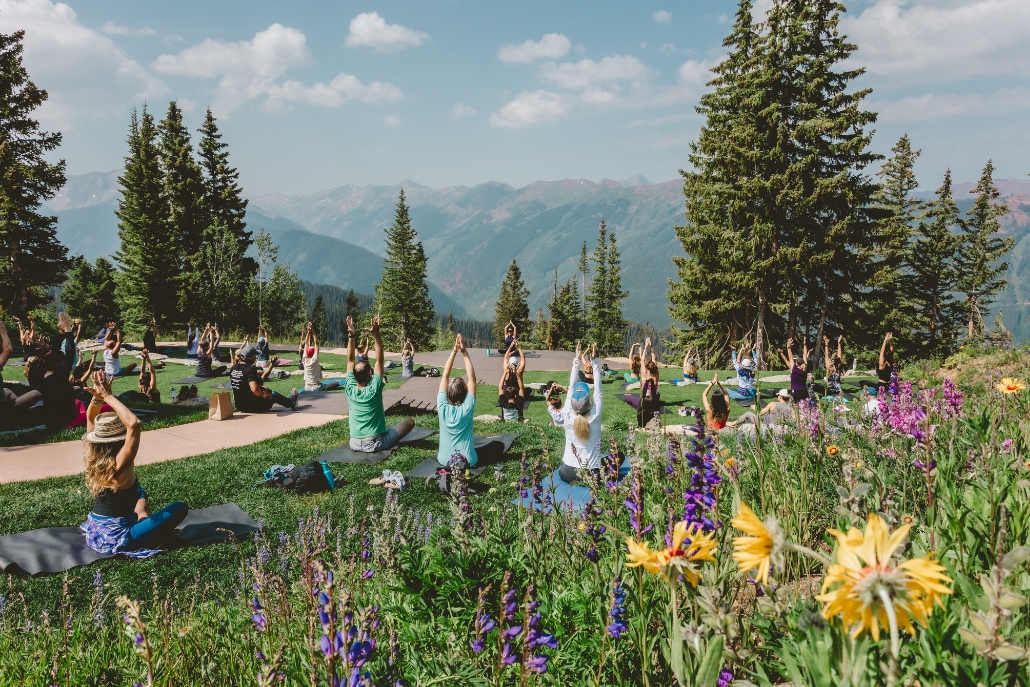A group of individuals doing yoga outdoors in the mountains, with vibrant flowers and trees, at Shakti Yoga Aspen.