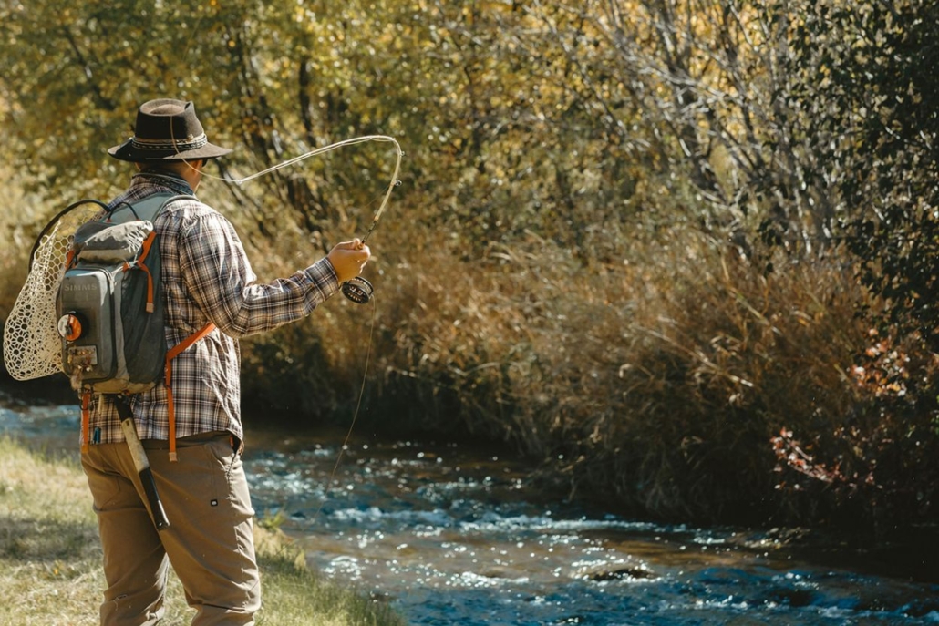 A man wearing a hat and carrying a backpack stands by a stream and ready for fly fishing, surrounded by the vibrant autumn colors of Aspen in October.