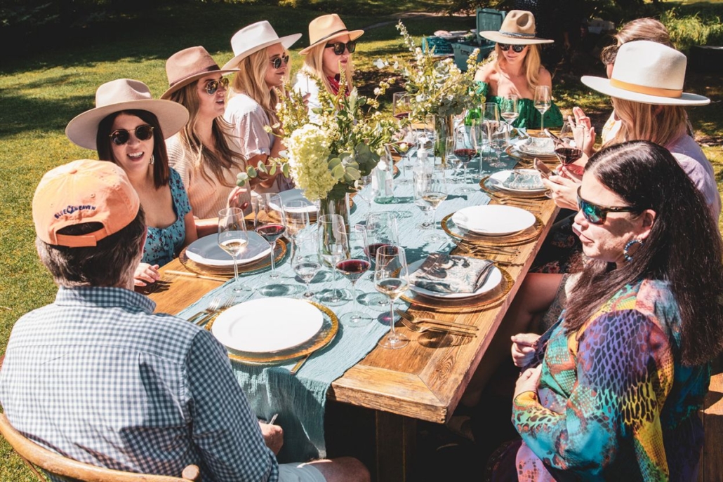 A group of individuals wearing hats gathers around a table during an aspen picnic, enjoying food and conversation.