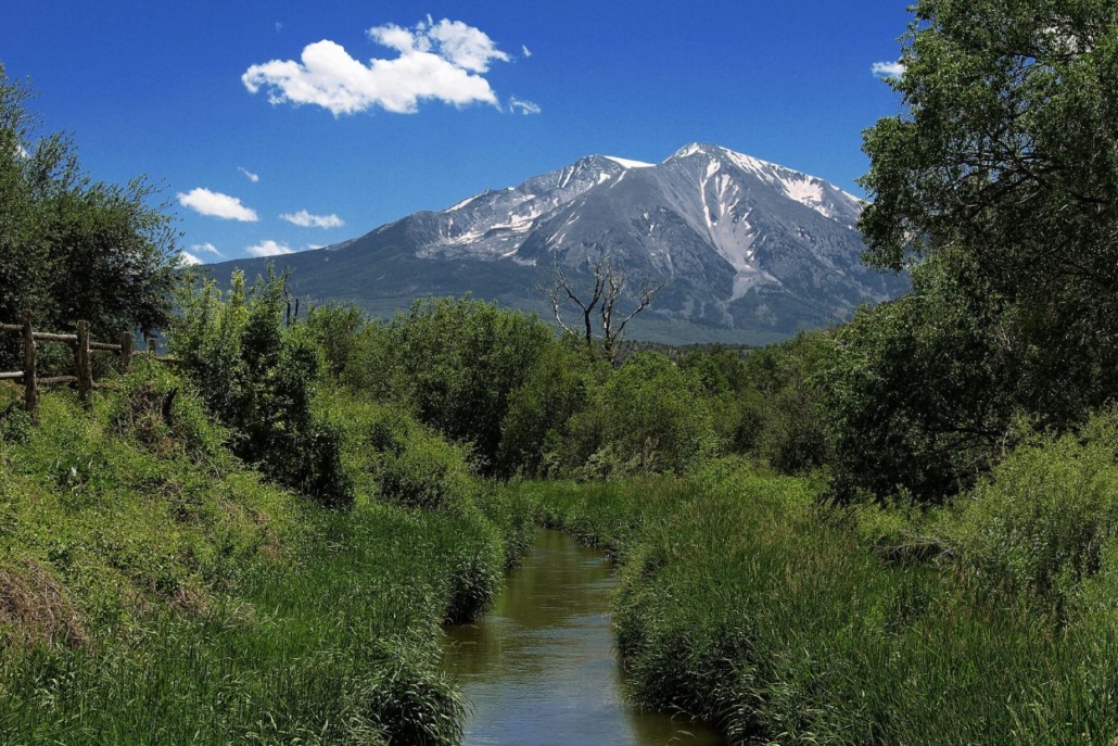 A tranquil stream meanders through a lush grassy field, framed by the stunning mountains of the Rio Grande Trail.