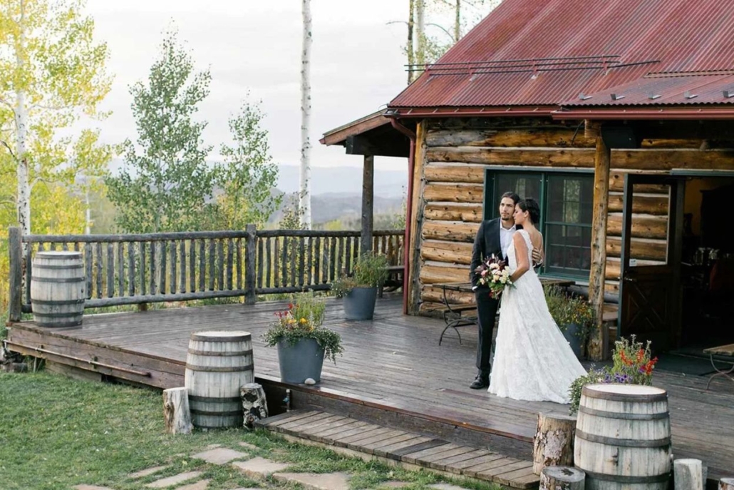  A bride and groom smile together on the porch of their rustic cabin, celebrating their aspen elopement in a beautiful setting.