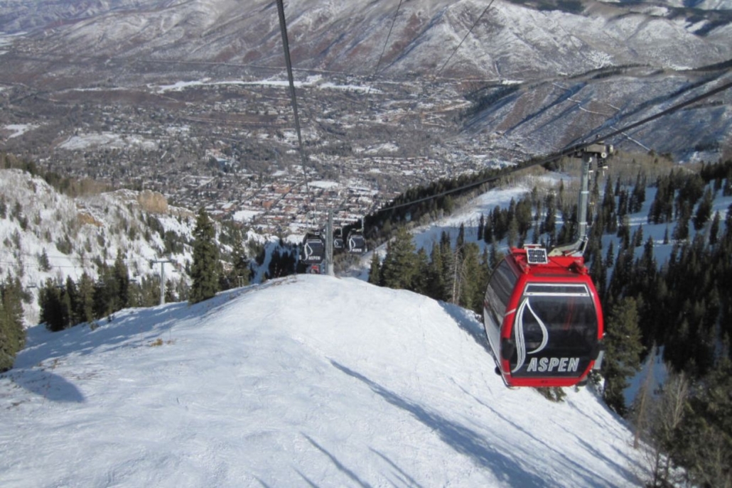 Red Aspen gondola lift ascending snowy mountain with scenic view of Aspen, Colorado, town and forested slopes in November.