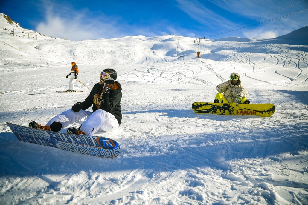 A snowboarder resting on the snow at Snowmass ski resort in Aspen, Colorado, with snow-covered mountains in the background.