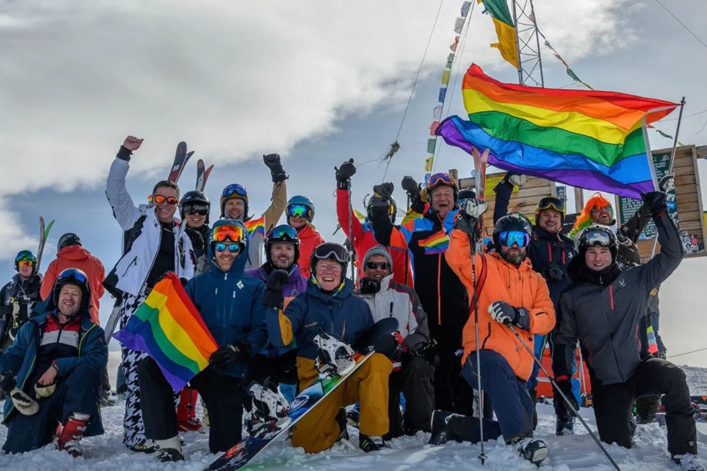 A diverse group of individuals proudly holding rainbow flags on a snowy mountain during Aspen Gay Ski Week.