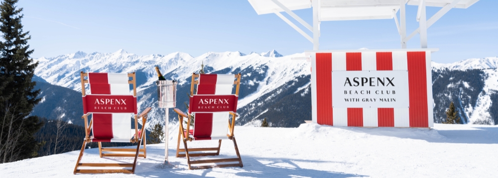 Aspen après-ski at AspenX Beach Club, featuring red lounge chairs, champagne on ice, and stunning mountain views against a clear blue sky.