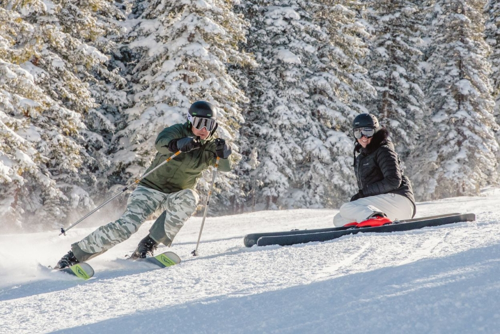 Two people enjoying Aspen in January, with one skiing and the other snowboarding on a snow-covered mountain surrounded by scenic, snow-draped trees.