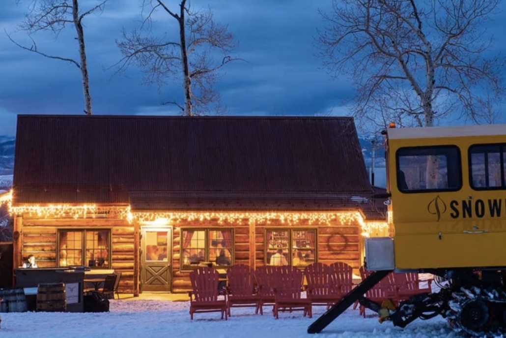 Rustic cabin adorned with string lights in a snowy landscape, setting the scene for a winter Aspen Snowcat Dinner experience under a twilight sky.