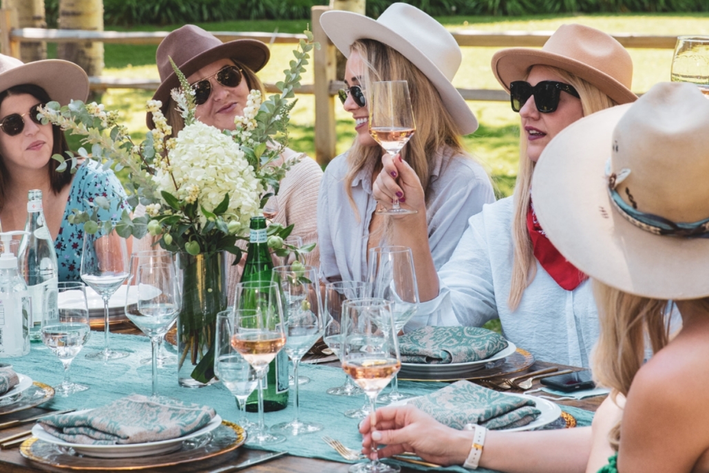 Women wearing hats and sunglasses seated at a table for a private wine tasting during an Aspen bachelorette celebration.