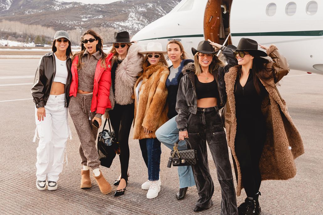 A cheerful group of women stands in front of an airplane, embodying the fashionable spirit of Aspen Style.