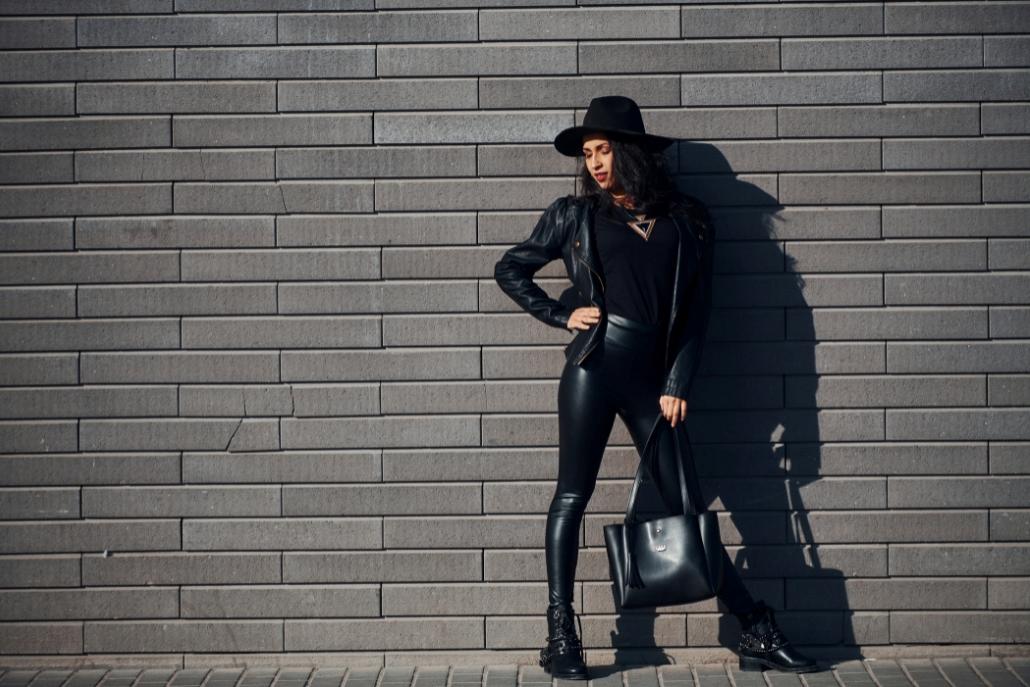 A stylish woman wearing black leather pants and a hat stands confidently against a rustic brick wall, showcasing Aspen Style.