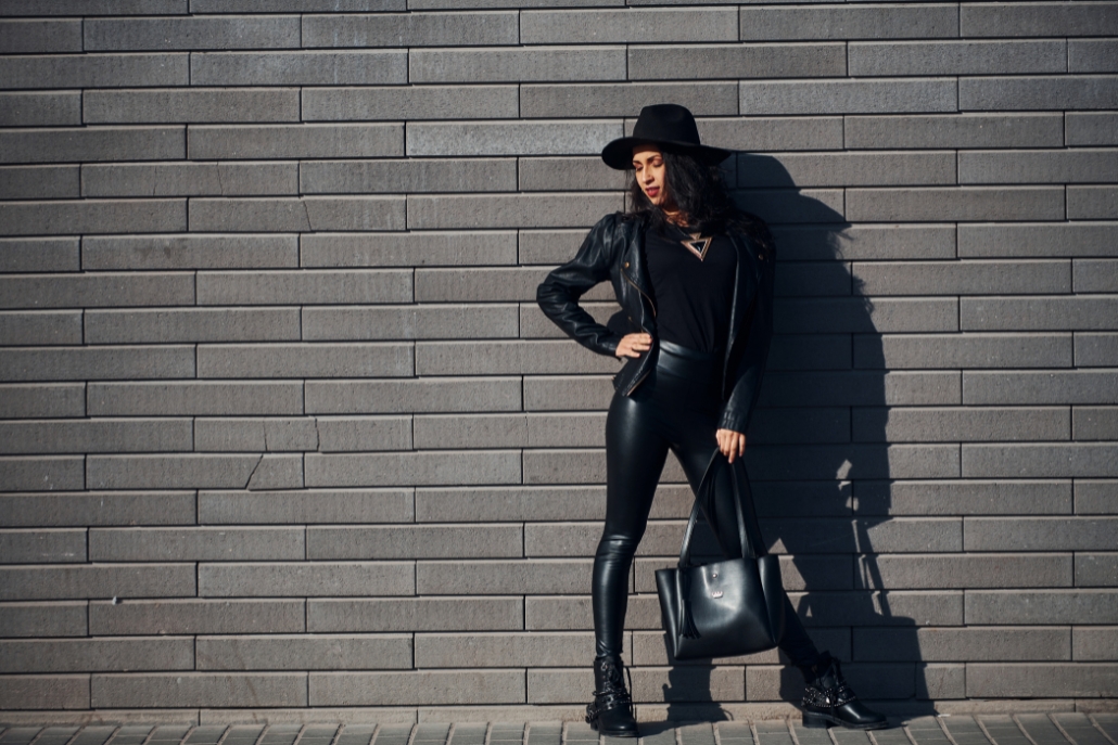 A stylish woman wearing black leather pants and a hat stands confidently against a rustic brick wall, showcasing Aspen Style.
