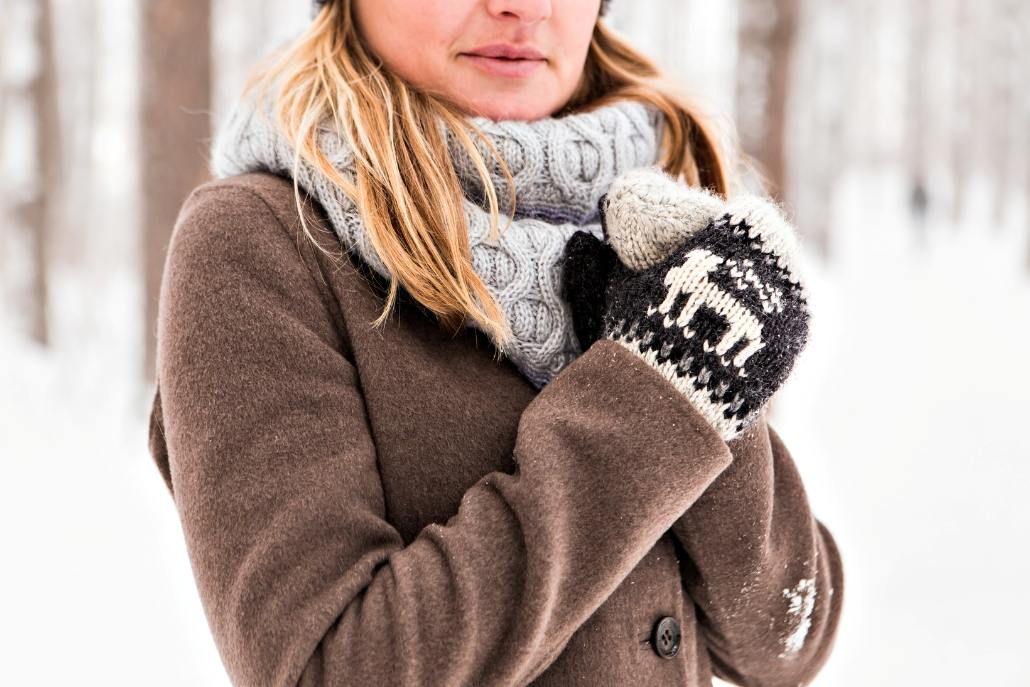 A woman in a winter coat and hat poses in the snow, representing Aspen Style's stylish warm scarves and accessories.