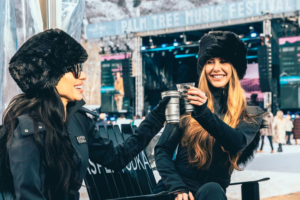 Two women wearing black Aspen style celebrate with a toast, donning black fur fats.