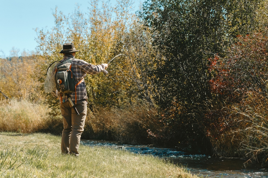 A man in a hat and plaid shirt stands on grass beside a stream, highlighting summer fly fishing activities in Aspen.