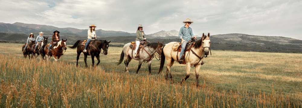 A group of people riding horses through a grassy field, enjoying Aspen summer activities in a scenic outdoor setting.