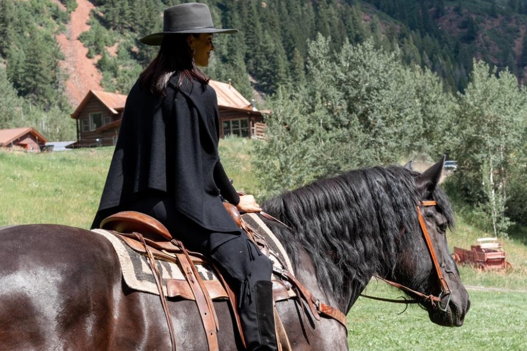A woman dressed in black rides a horse in the mountains, showcasing Aspen's summer horseback riding in Maroon Creek Valley.