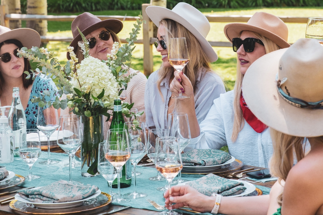 Women wearing hats and sunglasses seated at a table, participating in wine tasting at a private estate in Aspen.