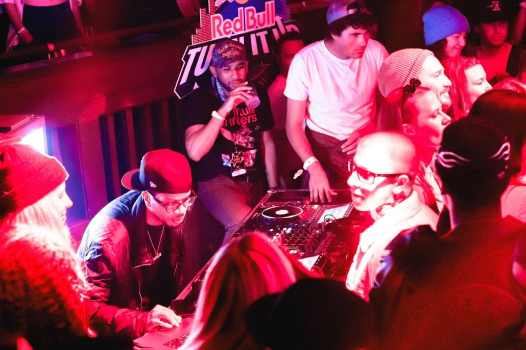 Crowd dancing near DJ booth under vibrant lights at a packed venue, energetic nightlife captured during a lively celebration.