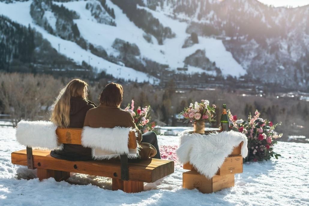 A couple sits on a wooden bench with white fur throws during an Aspen elopement, surrounded by snow-covered mountains and romantic floral arrangements, with champagne on a small wooden table.